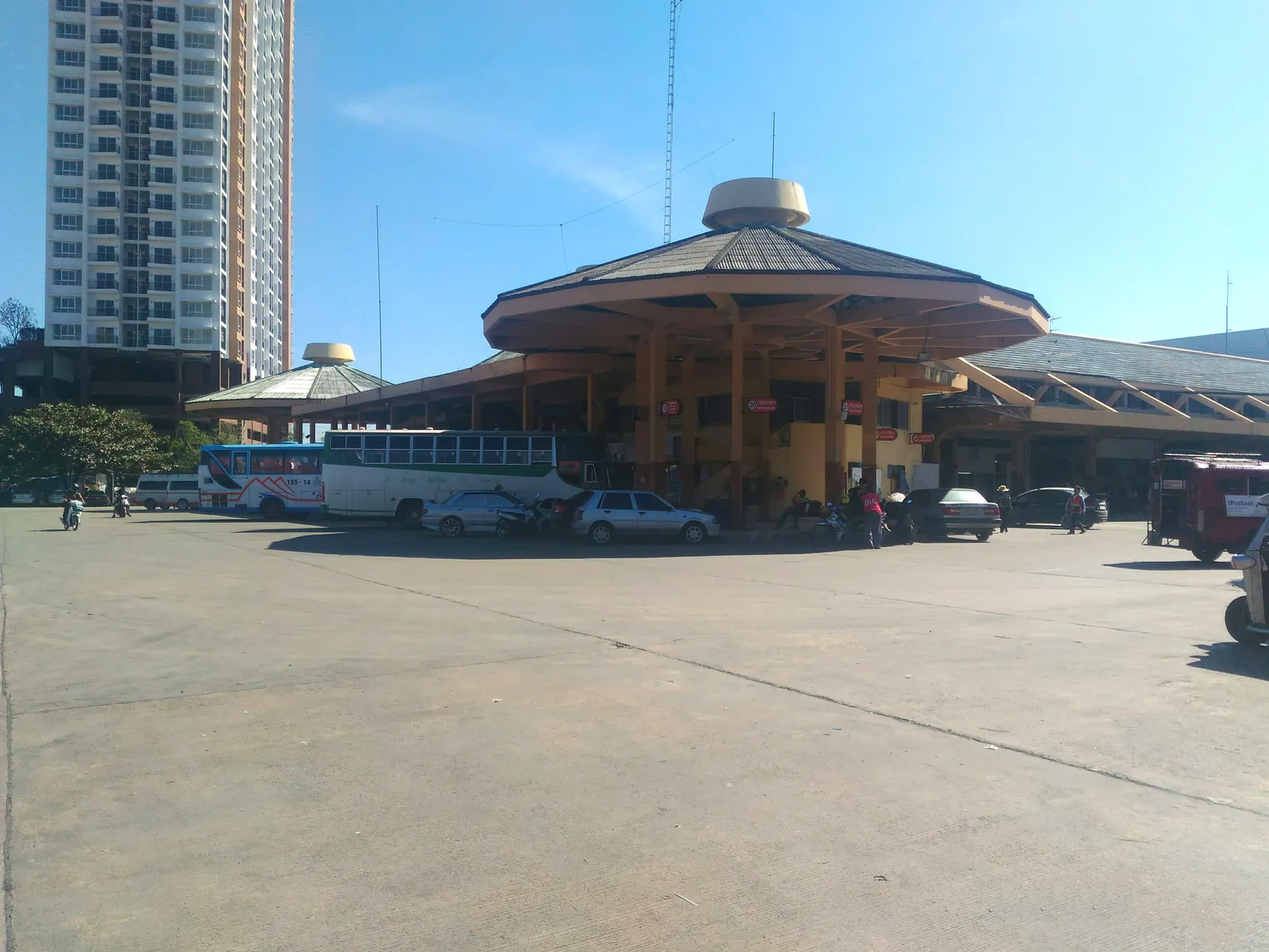 Octagonal roof over a bus bay at a station with parked buses, a tall apartment block behind and a wide concrete forecourt.