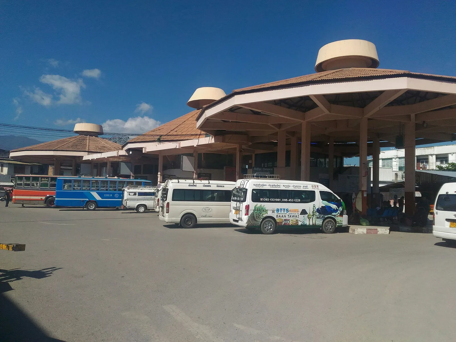 Bus station with several octagonal roofs, white minibuses parked in front and a blue bus at the left under a blue sky.