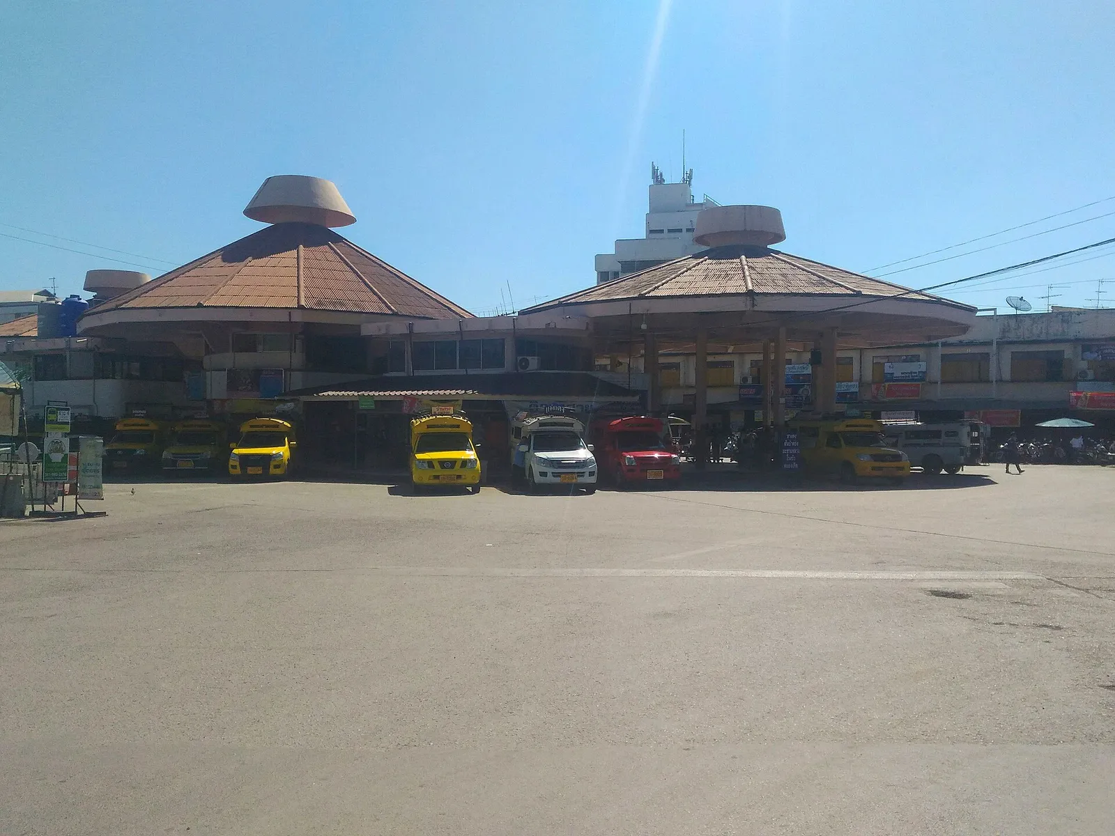 Row of parked yellow, red and white pickup-truck taxis in front of two octagonal-roofed station buildings.
