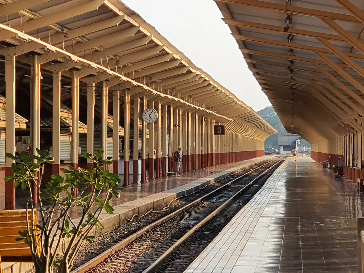 Long covered railway platform with roof supports, a track on one side and a bench and plant in the foreground.