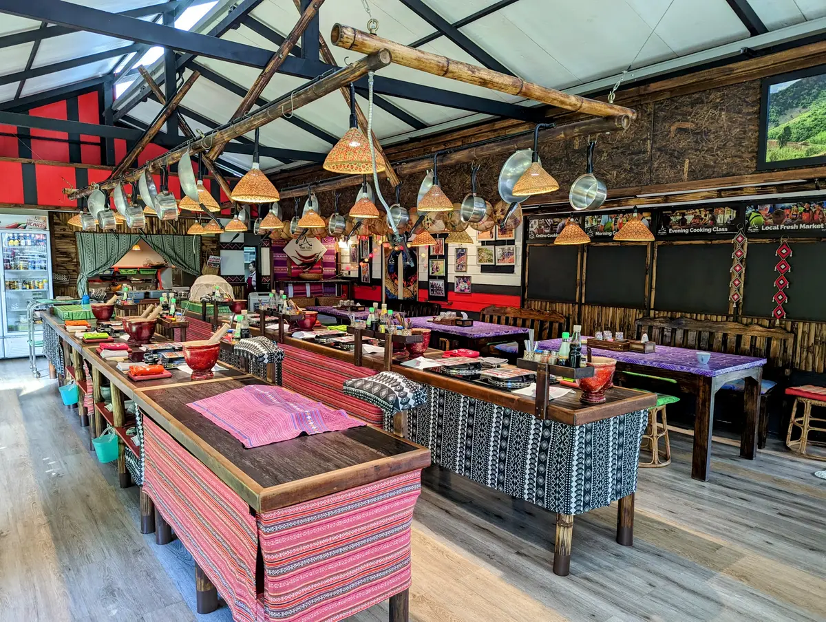 A long cooking hall with rows of wooden counters, woks and hanging lamps.