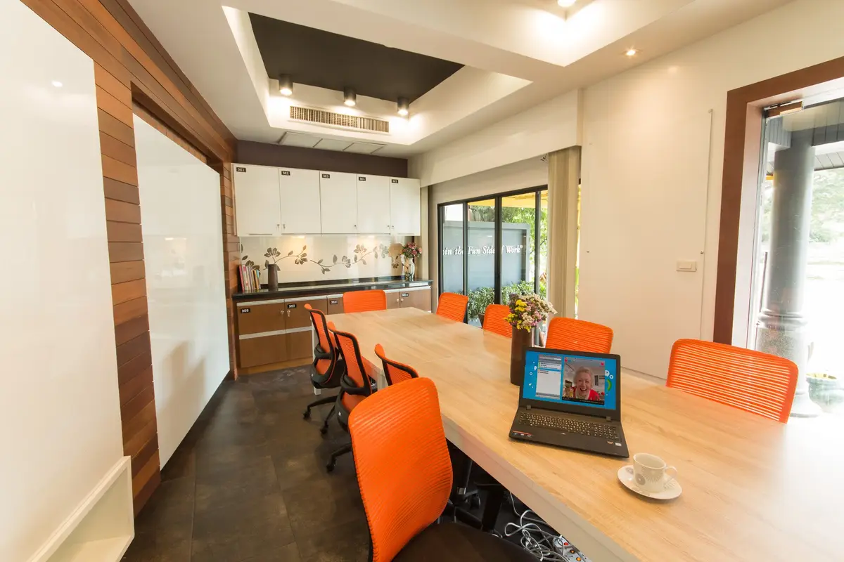 Shared work table with orange chairs, a laptop and a kitchen area in the background.