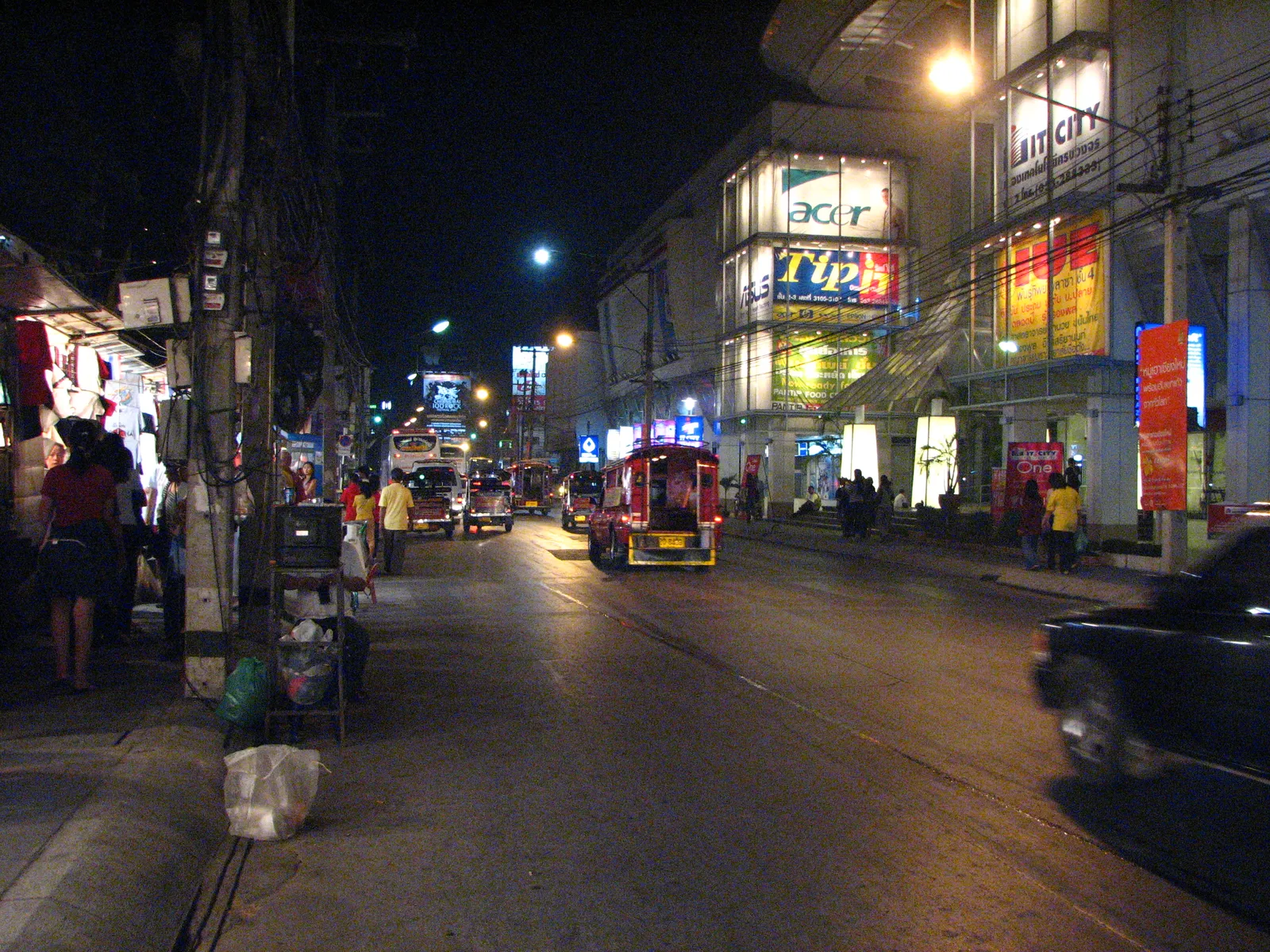 Wide street at night with a red songthaew and lit shop fronts.