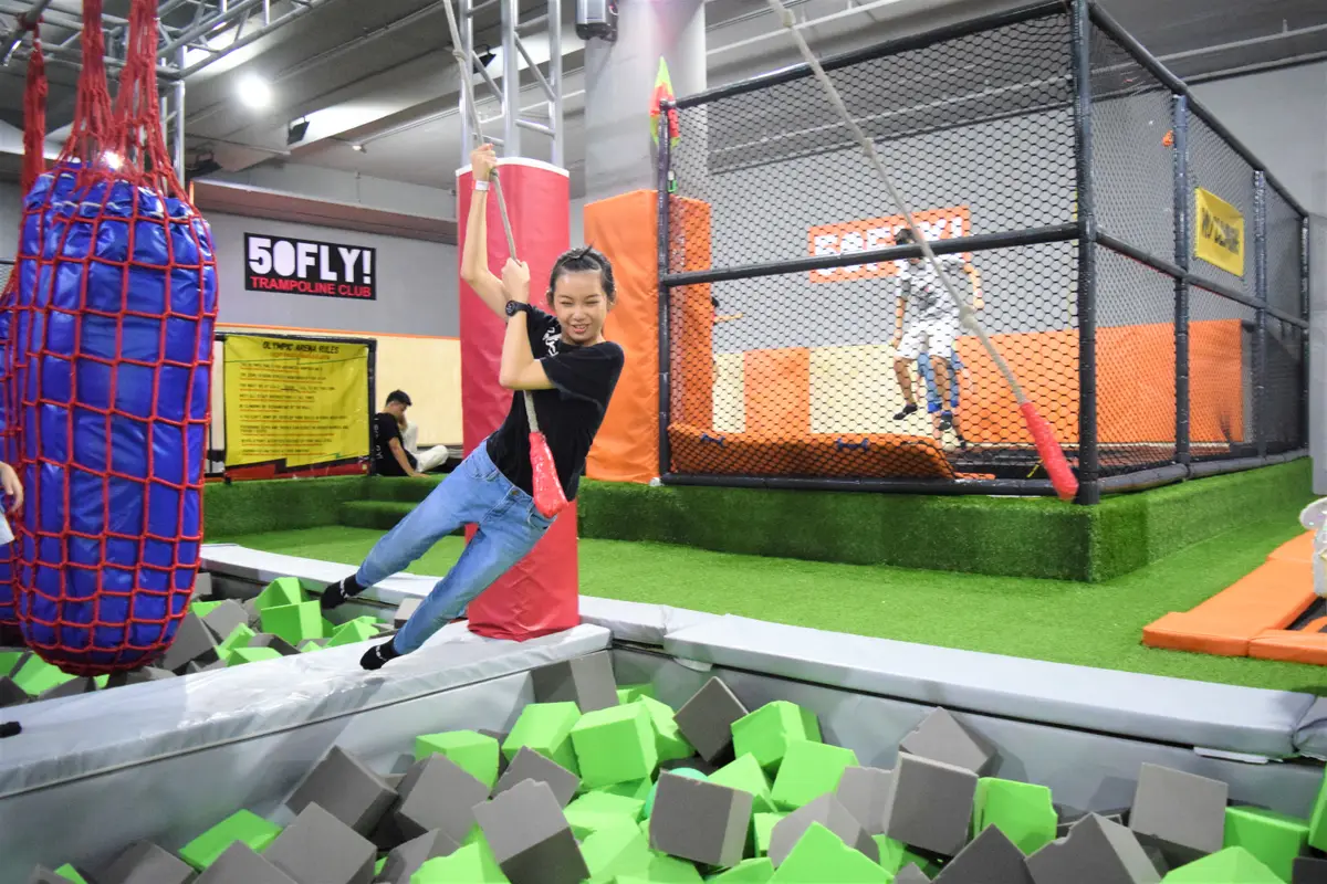 A girl swinging on a rope over a foam pit with a ninja course behind her.