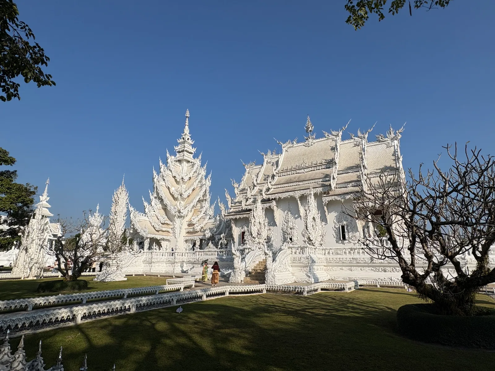 White temple halls with spired roofs behind a lawn and a bare tree