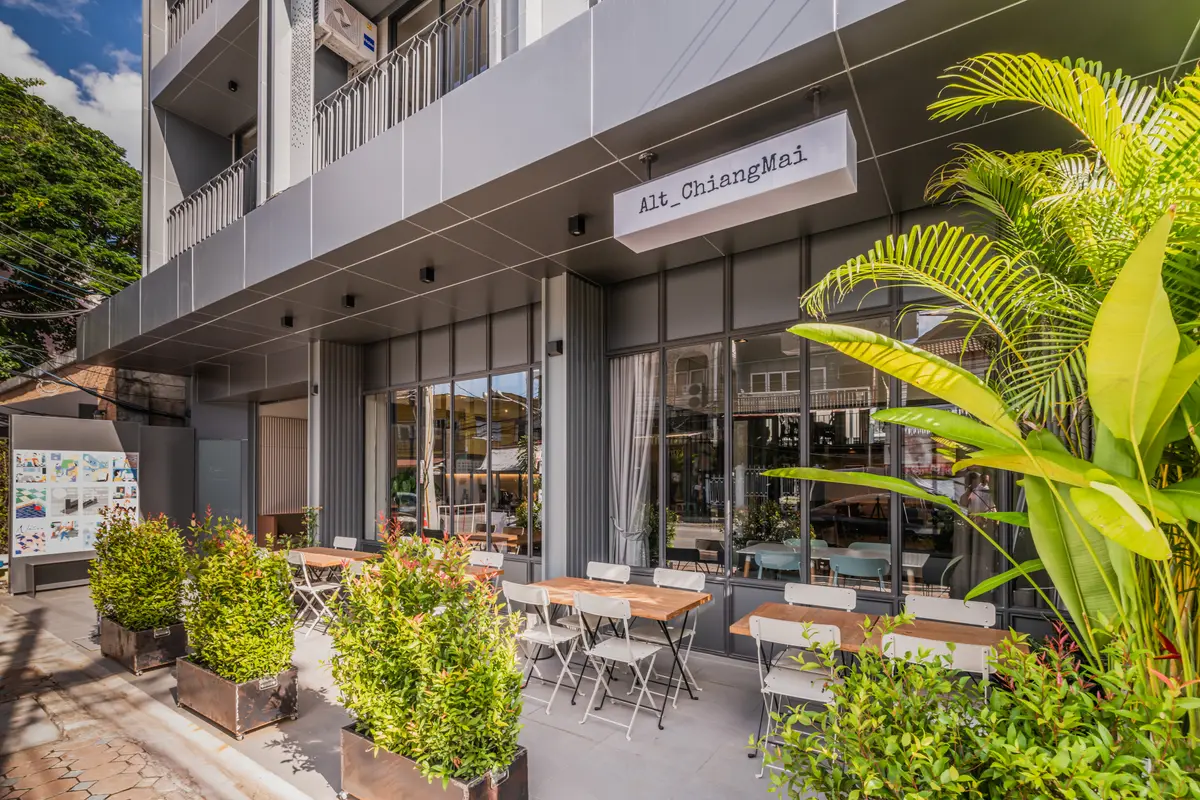 Grey building front with outdoor tables and chairs among potted plants and a hanging sign.