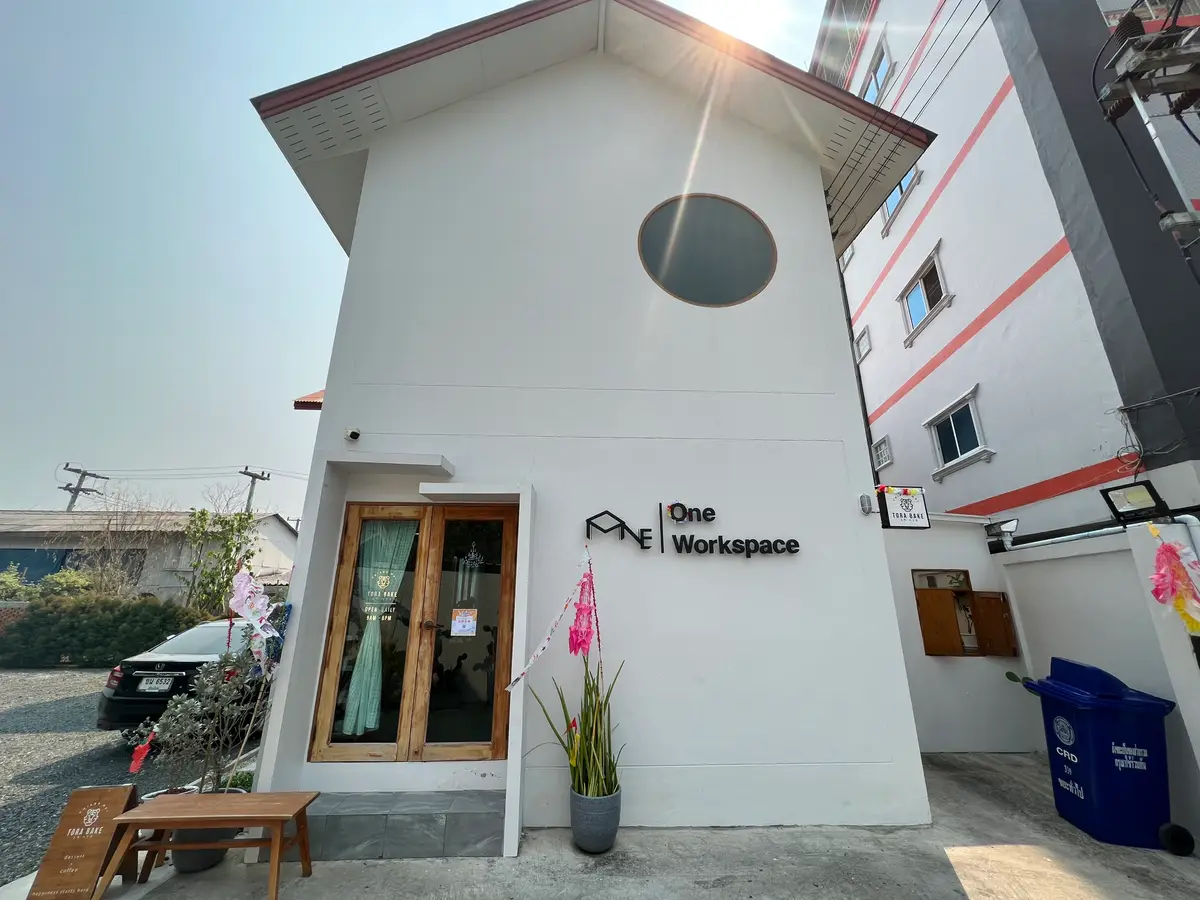 Small white building with a round window, wooden door and a sign reading One Workspace.