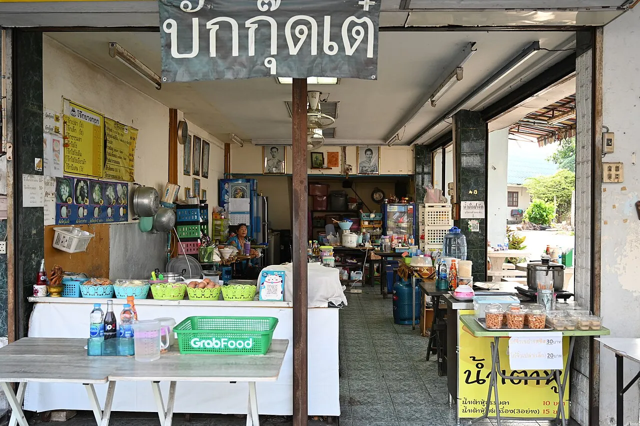 Open-fronted street eatery with a Thai sign, a counter of dishes and a green GrabFood tray on a table