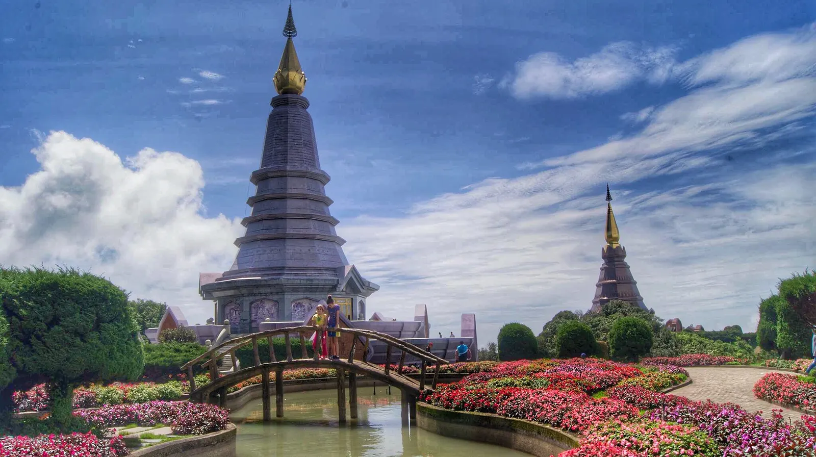 Two dark tiered pagodas with gold tips behind a flower garden and a wooden footbridge