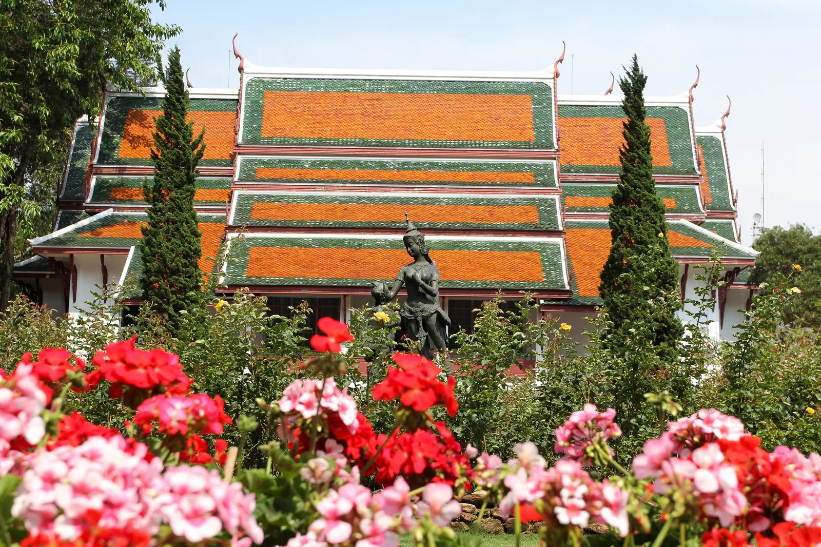 Palace building with green and orange tiled roofs behind flower beds and a statue