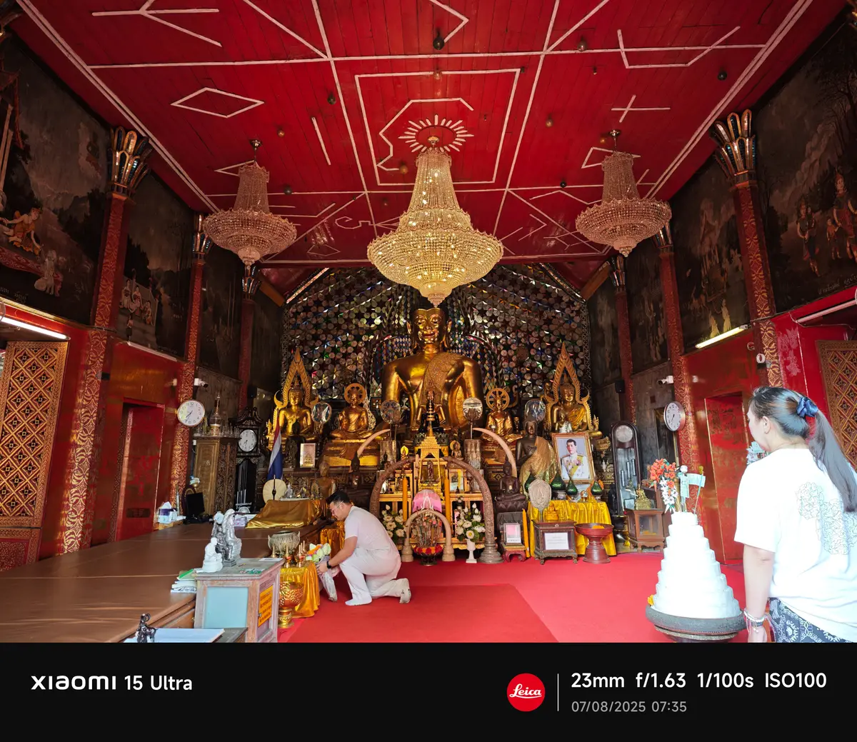 Gilded Buddha images on a tiered altar inside a hall with a red ceiling and chandeliers