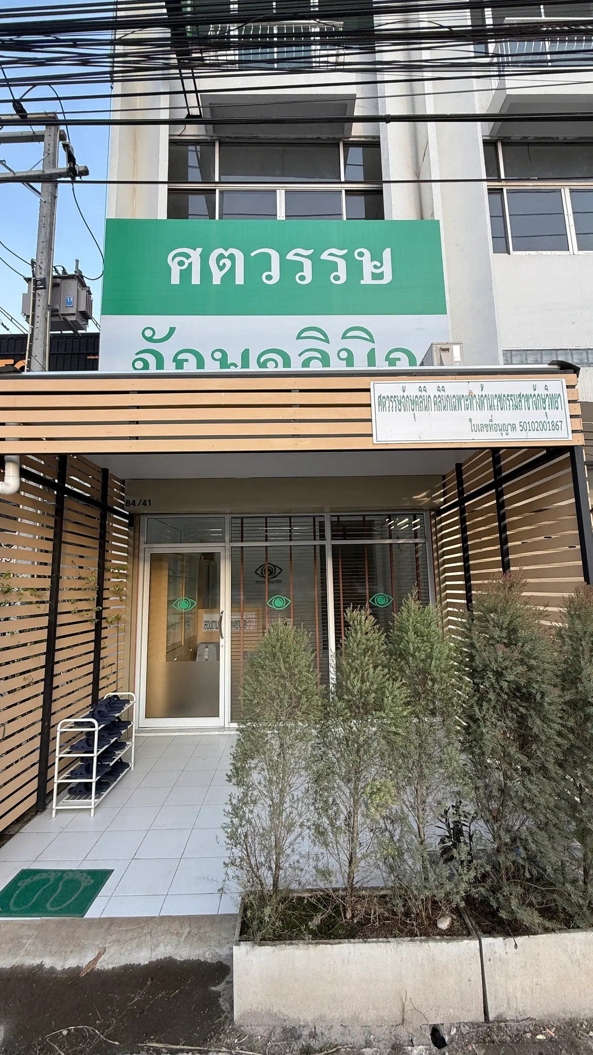 Glass front door of a small eye clinic with a green eye logo, wooden slat screens and shrubs beside the entrance