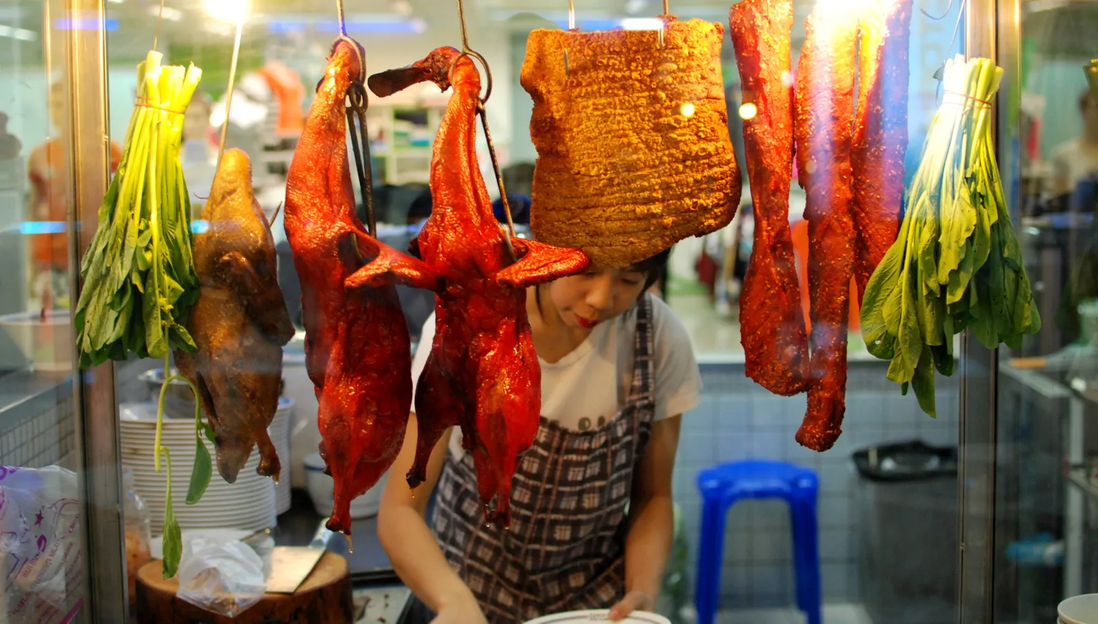 Roast ducks and slabs of crispy pork hanging in a food stall window beside green vegetables.