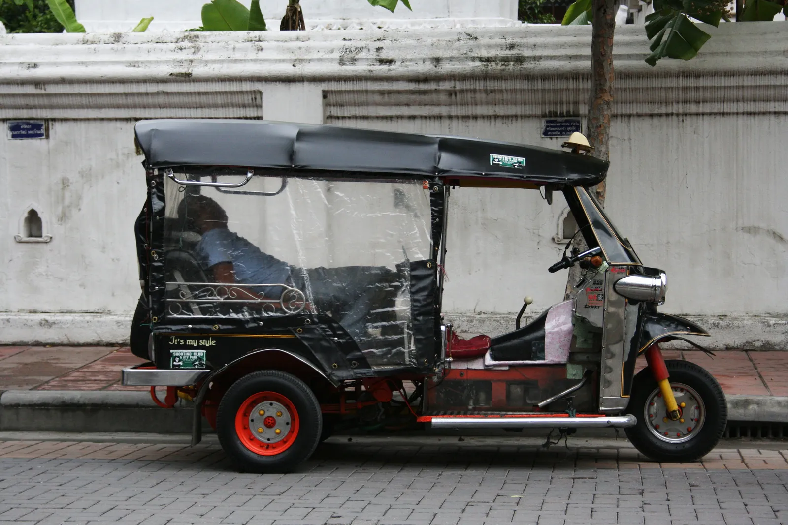 Black three-wheeled tuk-tuk with a clear plastic side curtain parked beside a white wall