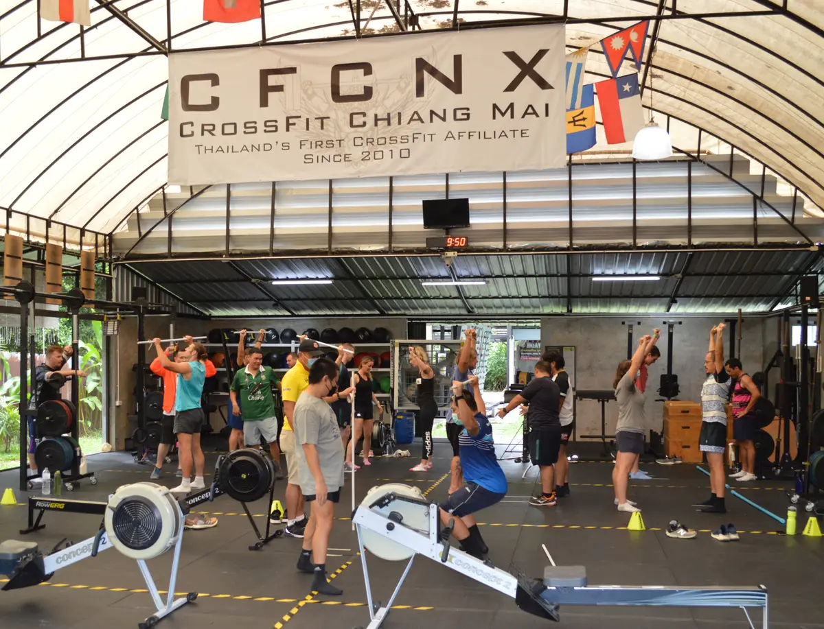 Group of people doing a class under a curved roof with rowing machines in the foreground.