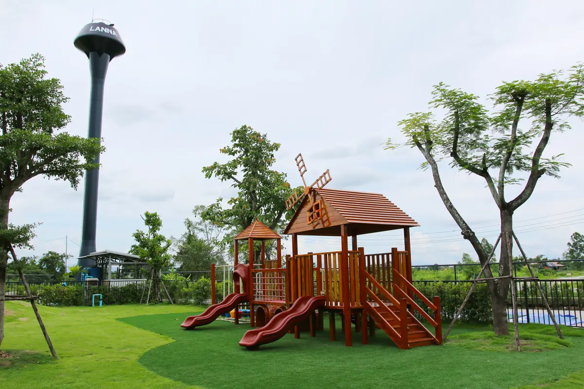 Wooden play tower with slides on a lawn, with a water tower and trees behind