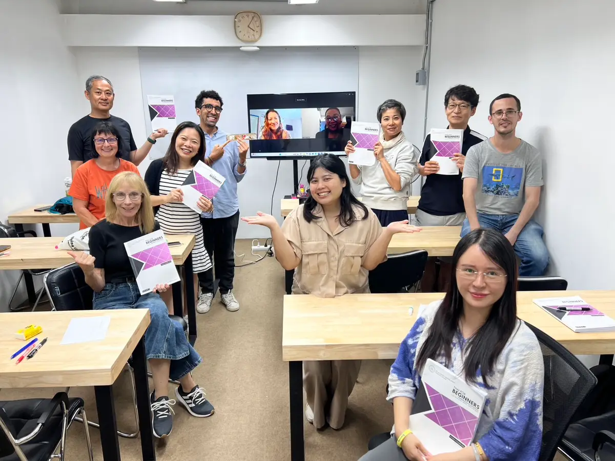 Students at desks in a bright classroom holding course books and smiling at the camera