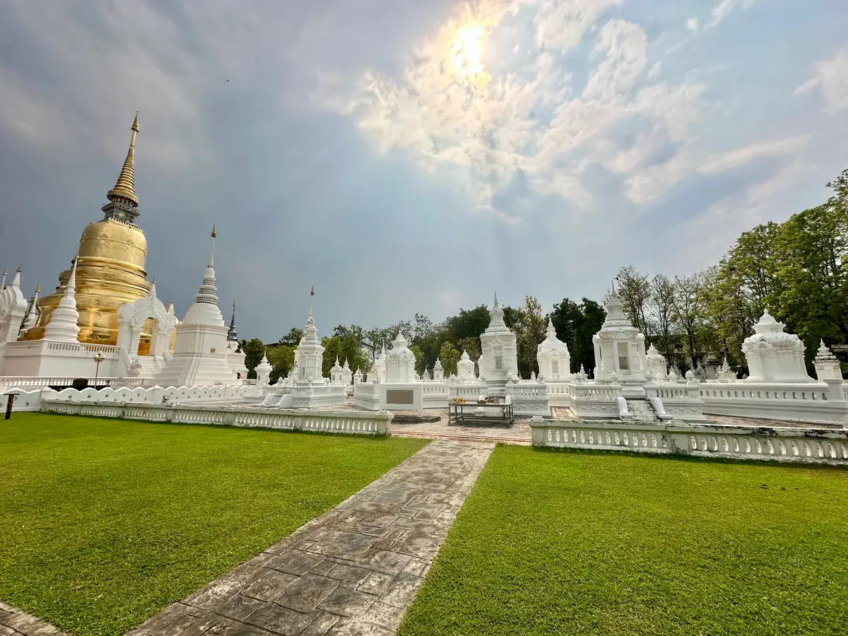 Rows of white chedis beyond a green lawn, with a gold chedi at the left under a cloudy sky