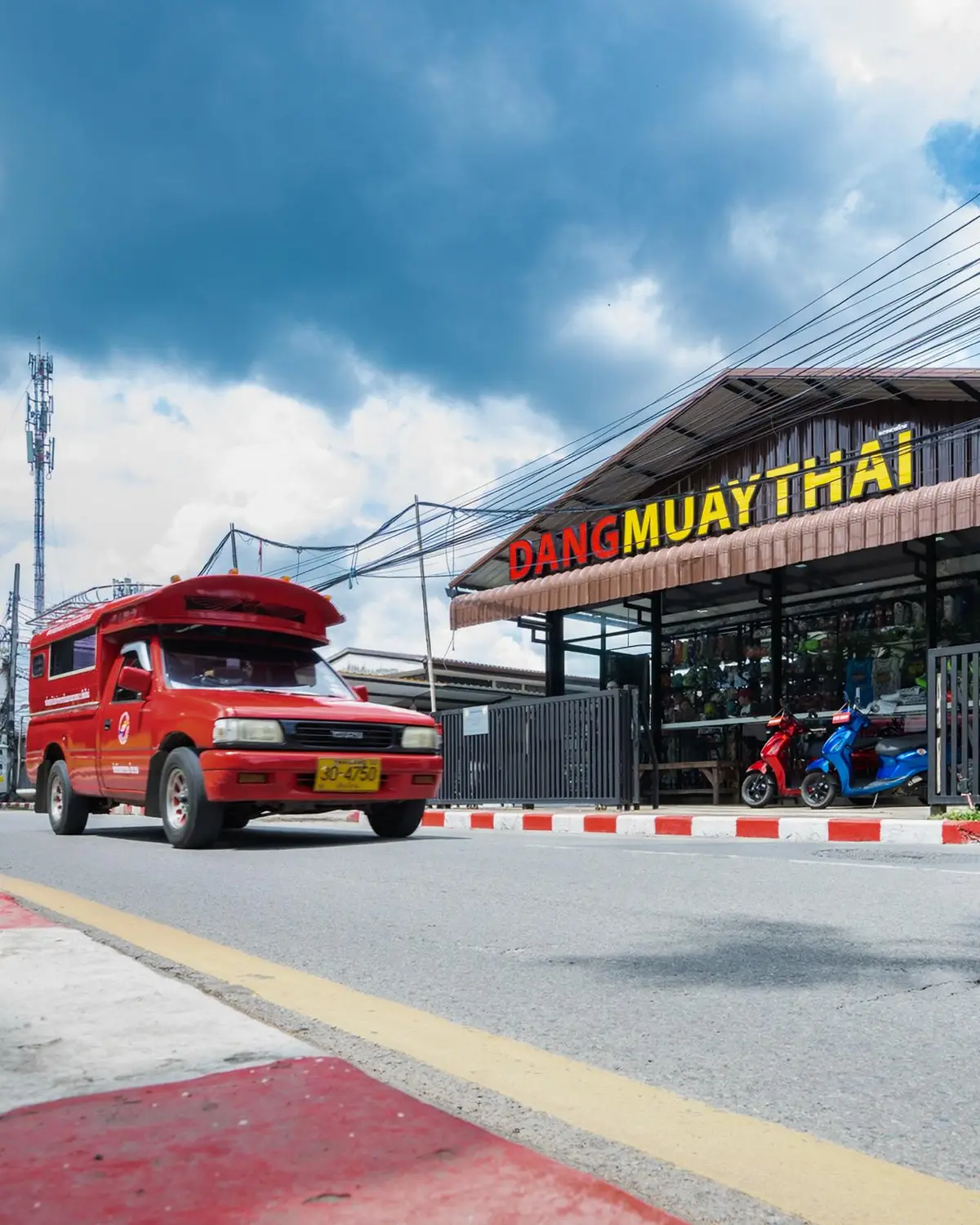 Shop front with a yellow sign reading Dang Muay Thai and a red songthaew passing on the street.