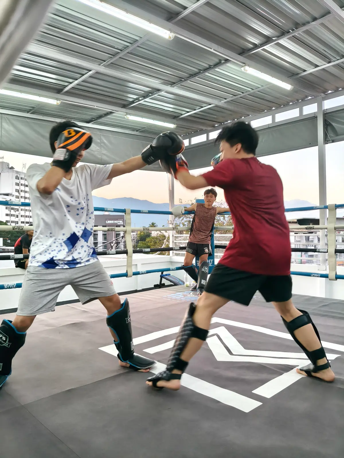 Two trainees in shin guards sparring on a rooftop ring with city buildings behind.