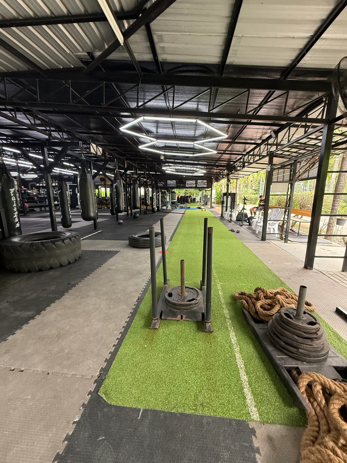 Long covered training area with heavy bags, tyres, a weighted sled and a strip of green turf.