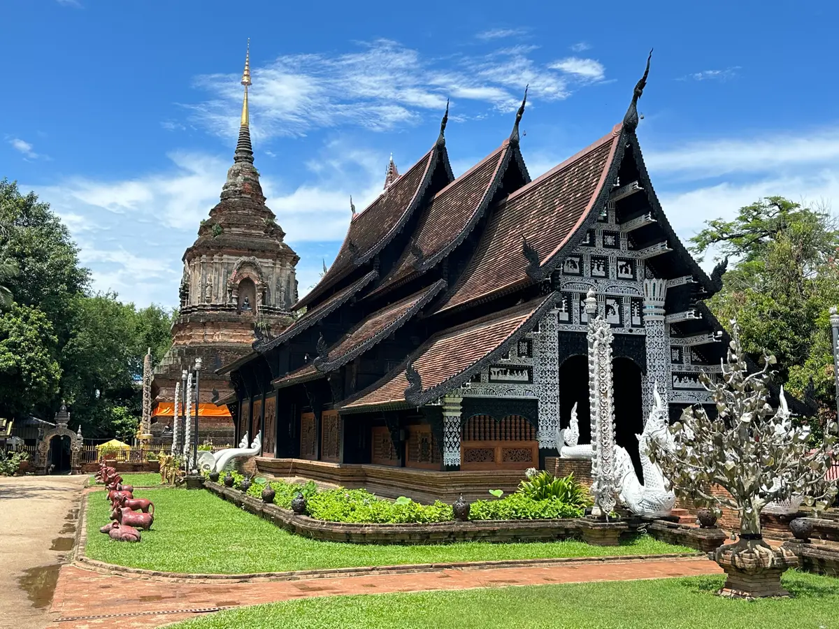 Dark wooden hall with steep stacked roofs and a brick chedi behind it, with a green lawn in front