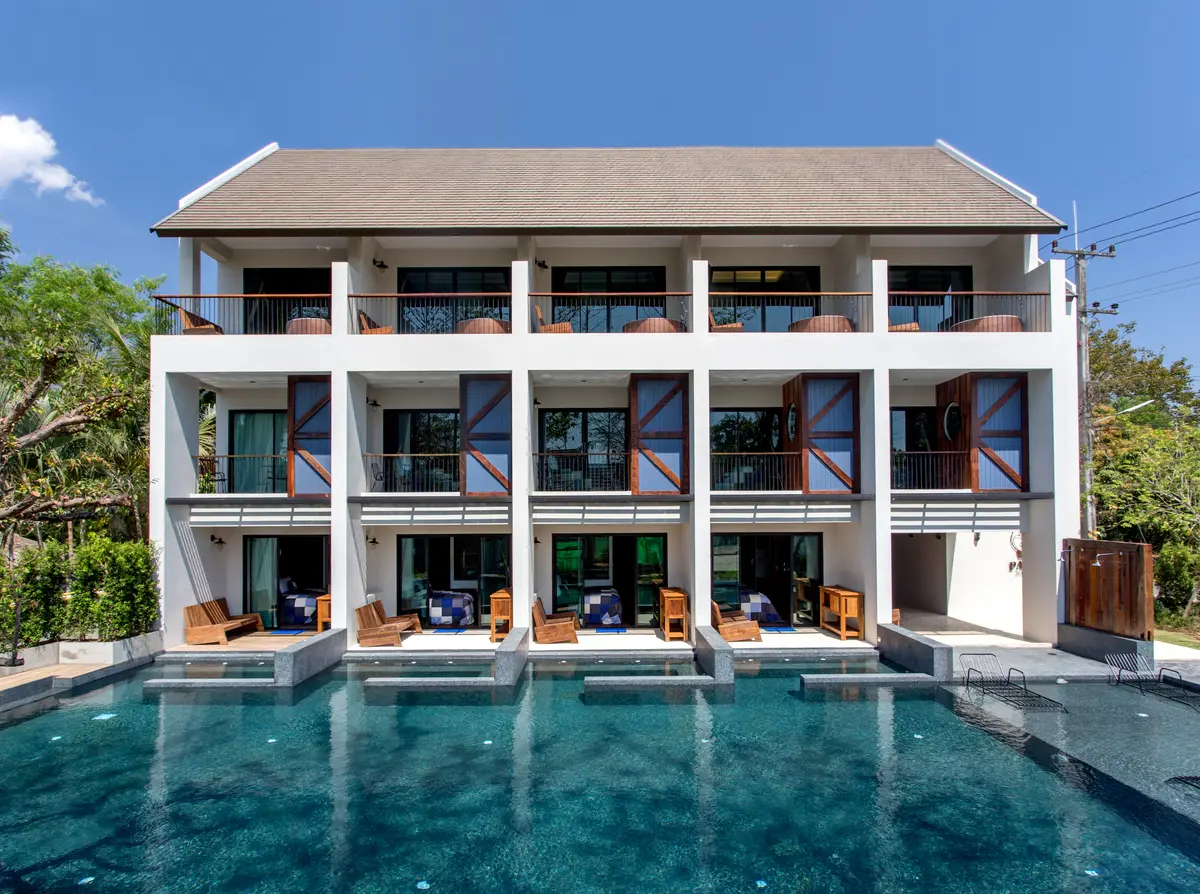 Two-storey white hotel building with balconies behind a long blue swimming pool