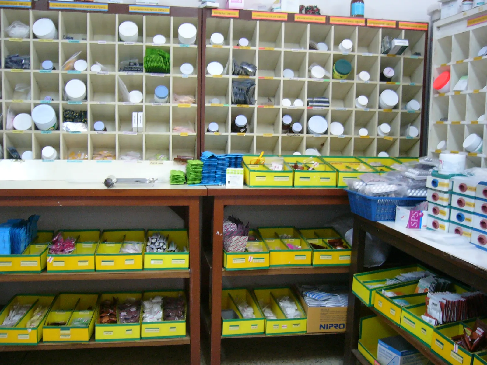 Wall of pigeonholes holding medicine tubs above yellow trays of sorted tablets in a hospital pharmacy.