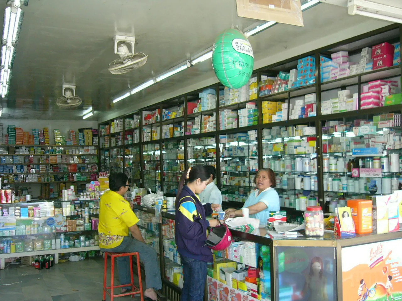 Inside a Thai pharmacy, shelves of boxed medicines behind a counter where a customer talks to a woman serving.