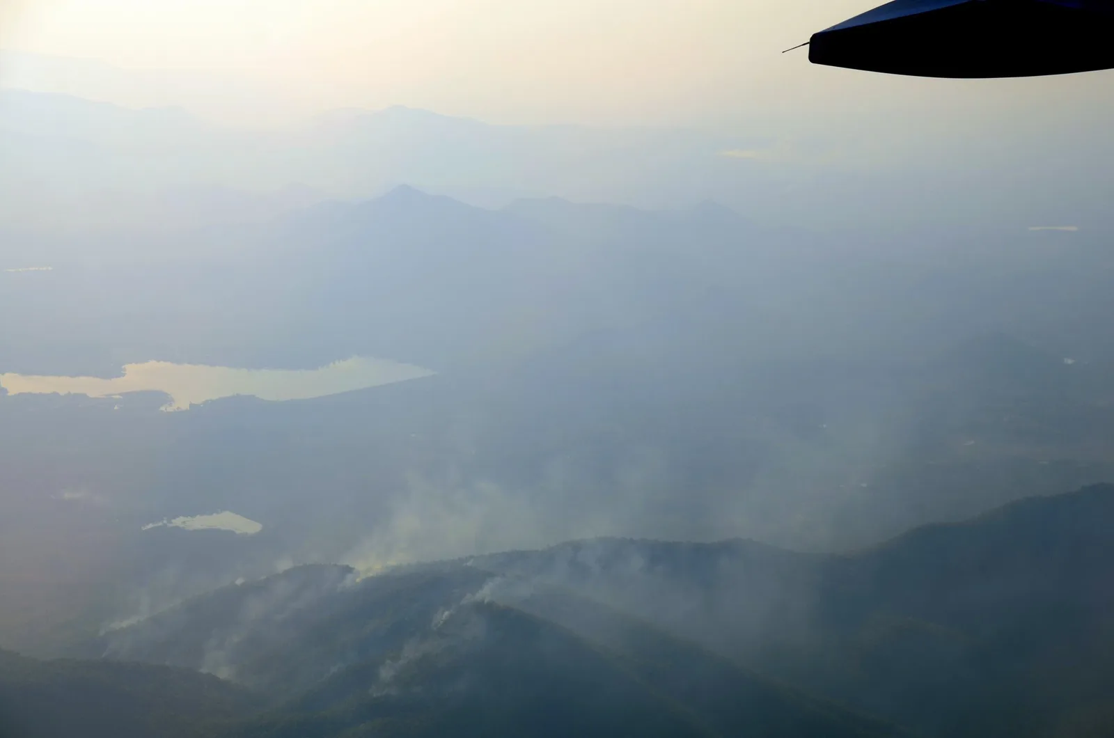 Aerial view of grey smoke rising from several fires on forested mountain slopes, with a lake and hazy ridges beyond.