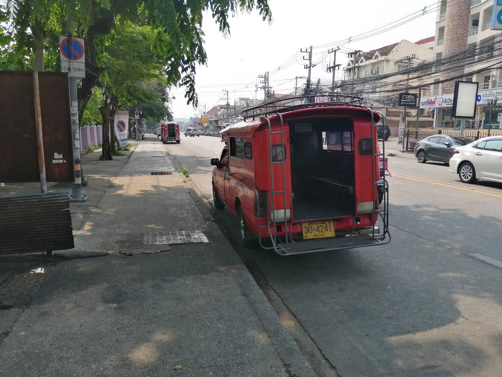 Red covered pickup truck seen from behind parked at the kerb of a tree-lined street