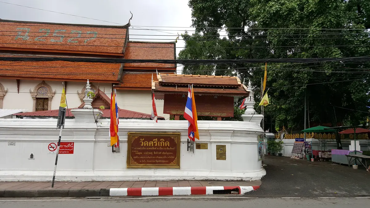 A white temple wall with a wooden name plaque, flags and a red-tiled roof behind.