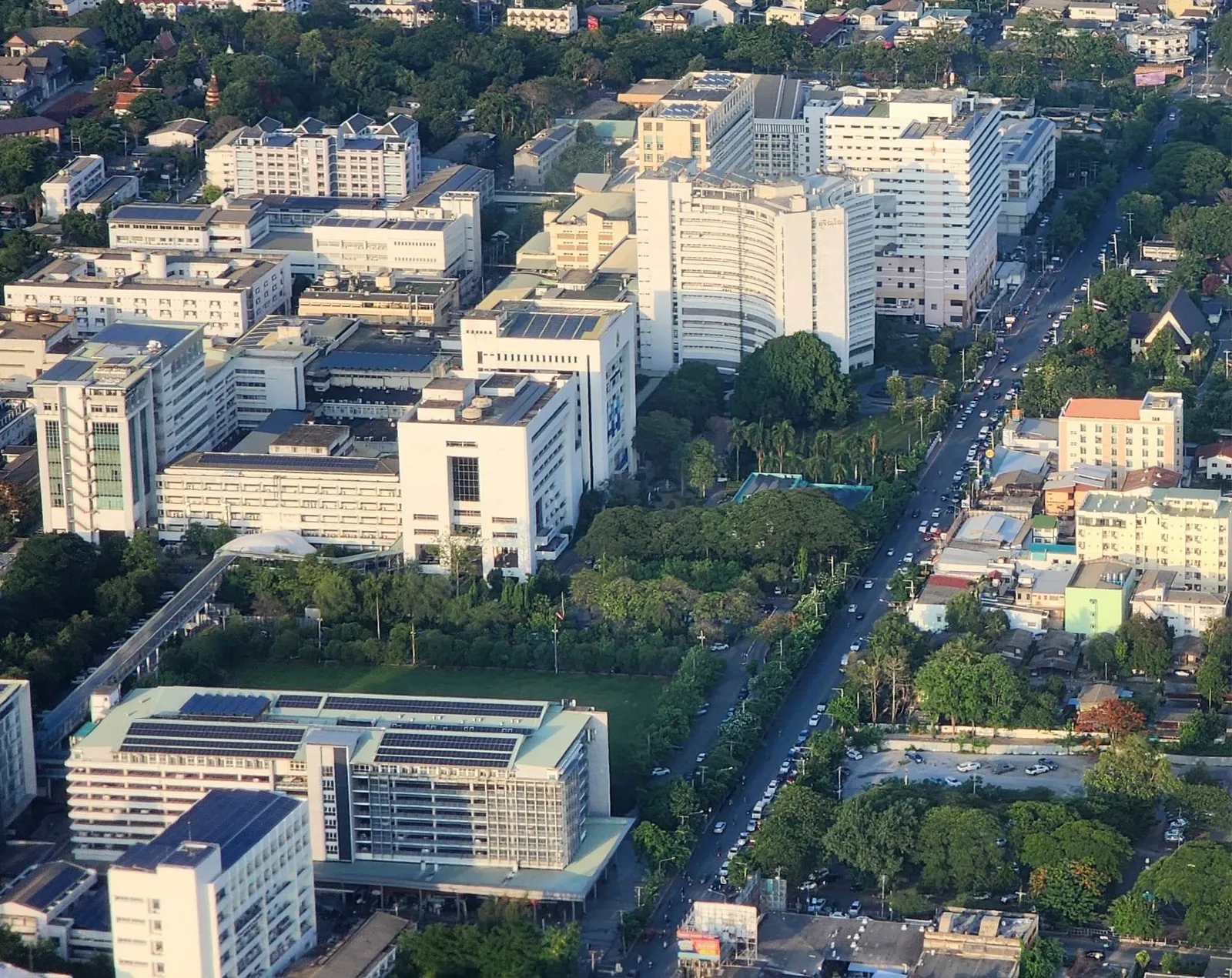 Aerial view of a dense hospital campus with several white multi-storey buildings beside a tree-lined road.