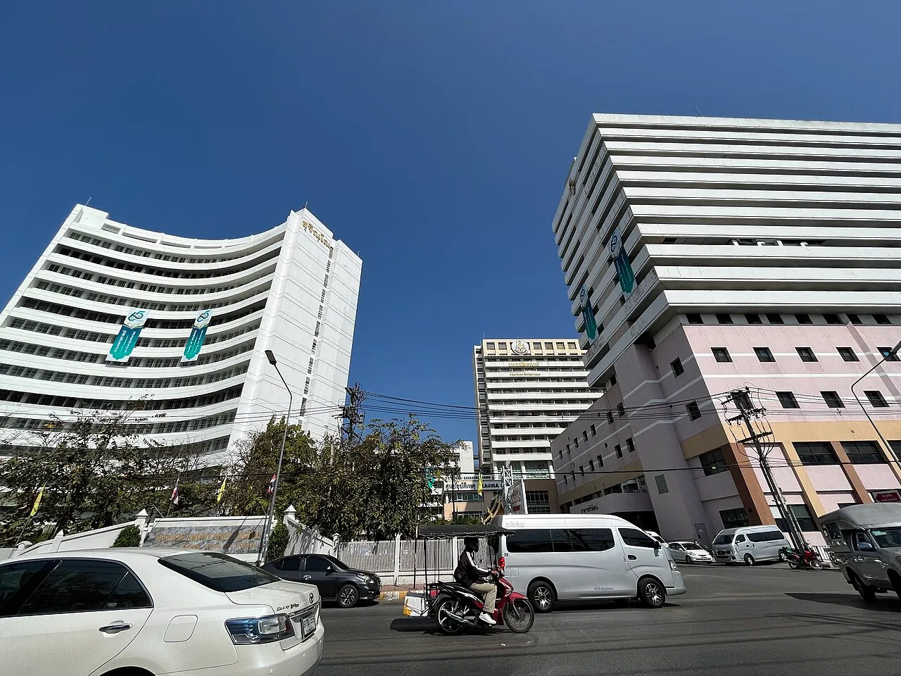 Curved white hospital tower and a pink building beside a street with a white van and a motorbike.