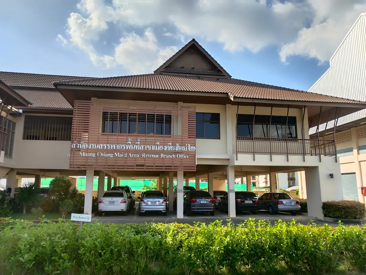 Low Thai-style government building with a brown roof and parked cars in front of a trimmed hedge.