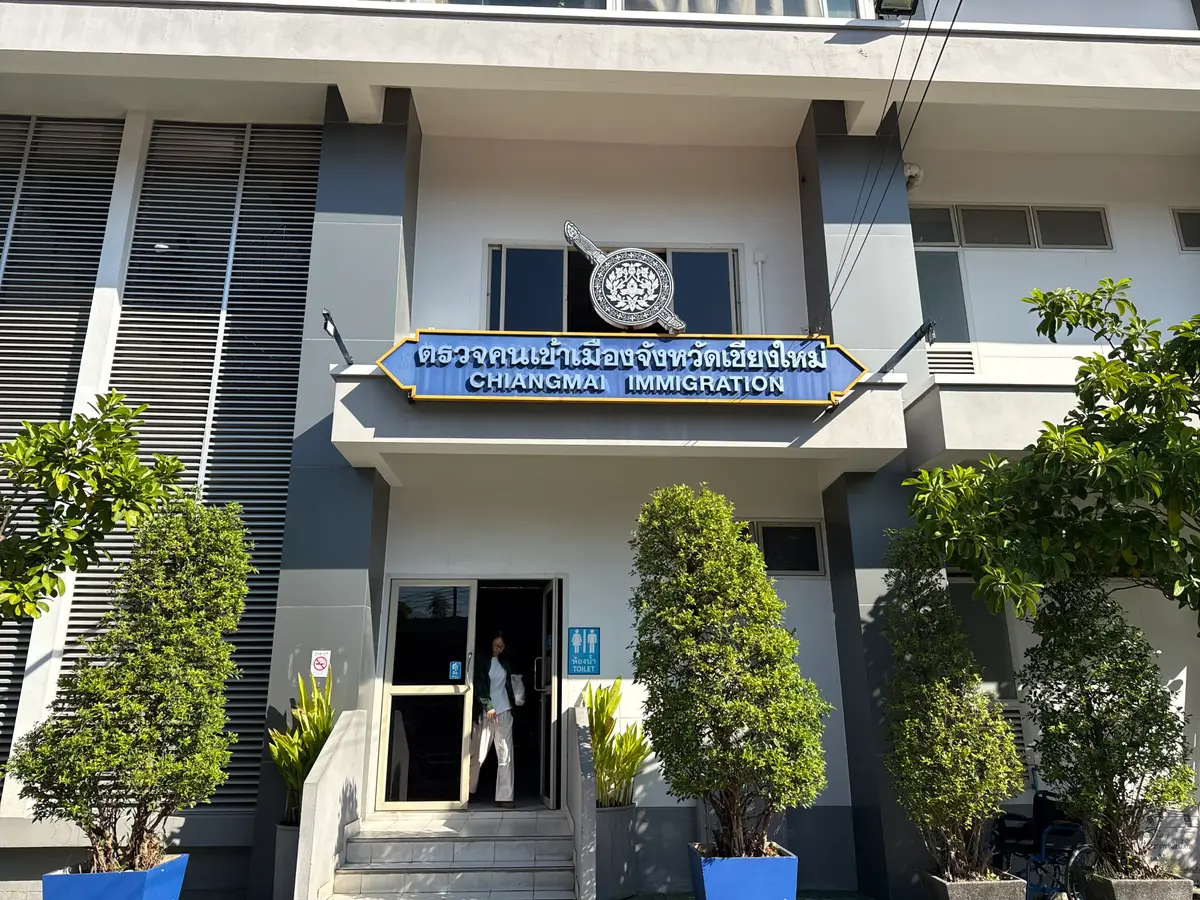 Entrance of the Chiang Mai Immigration office, a two-storey building with a blue name sign and trimmed shrubs in front.