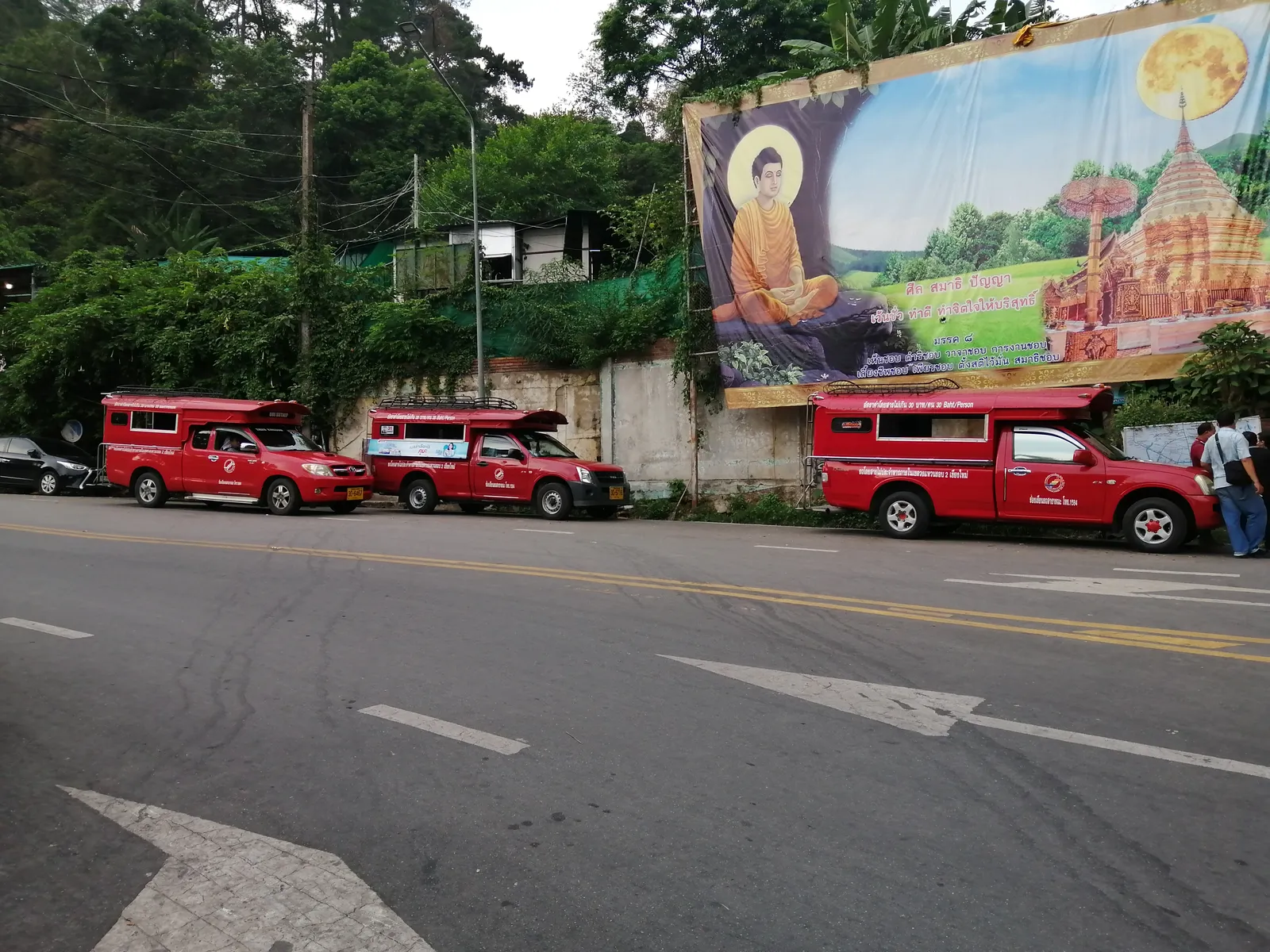Three red pickup trucks lined up on a road below a large painted billboard of a temple
