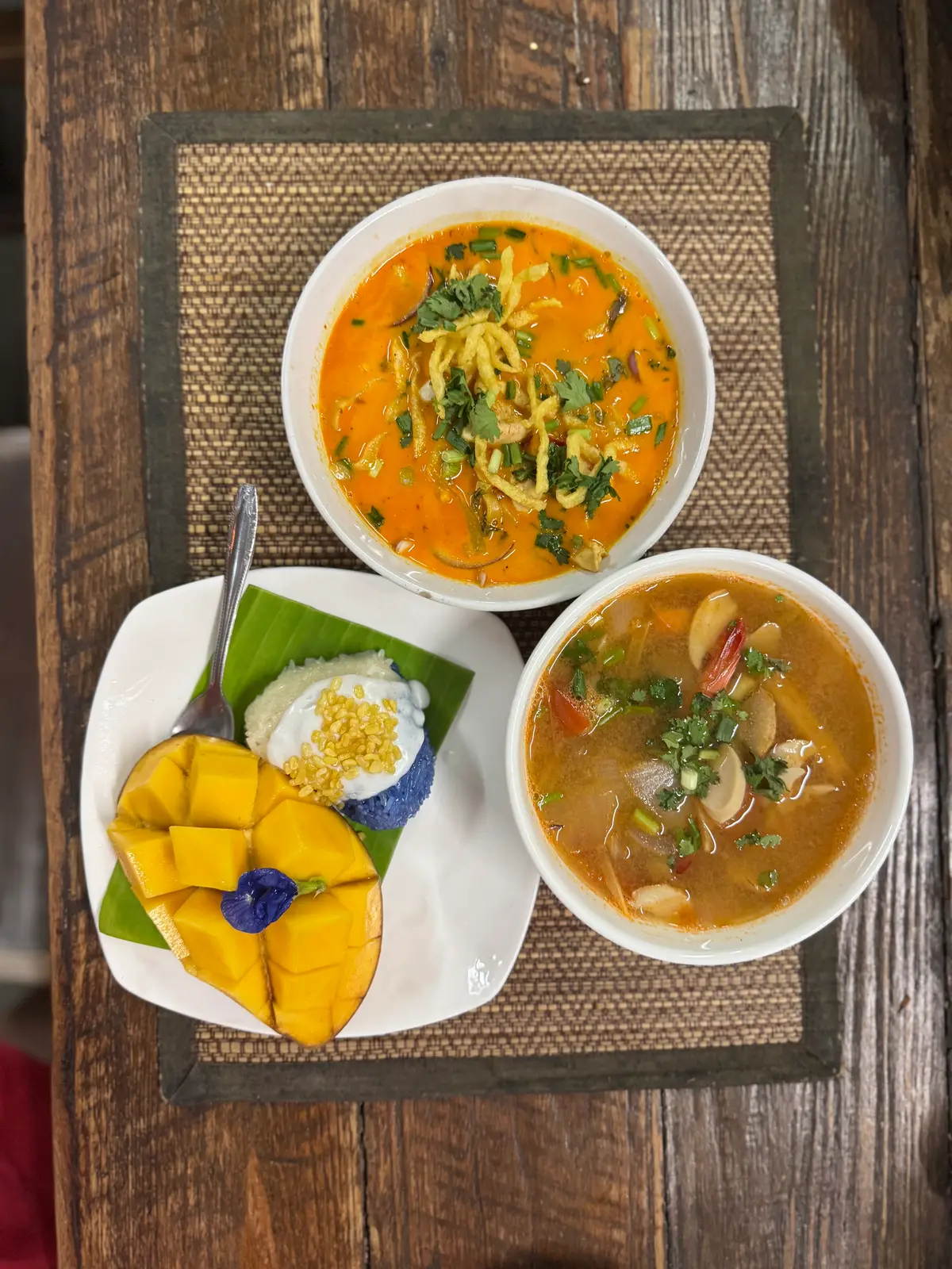Bowls of orange curry noodle soup and hot and sour soup beside mango with sticky rice on a banana leaf