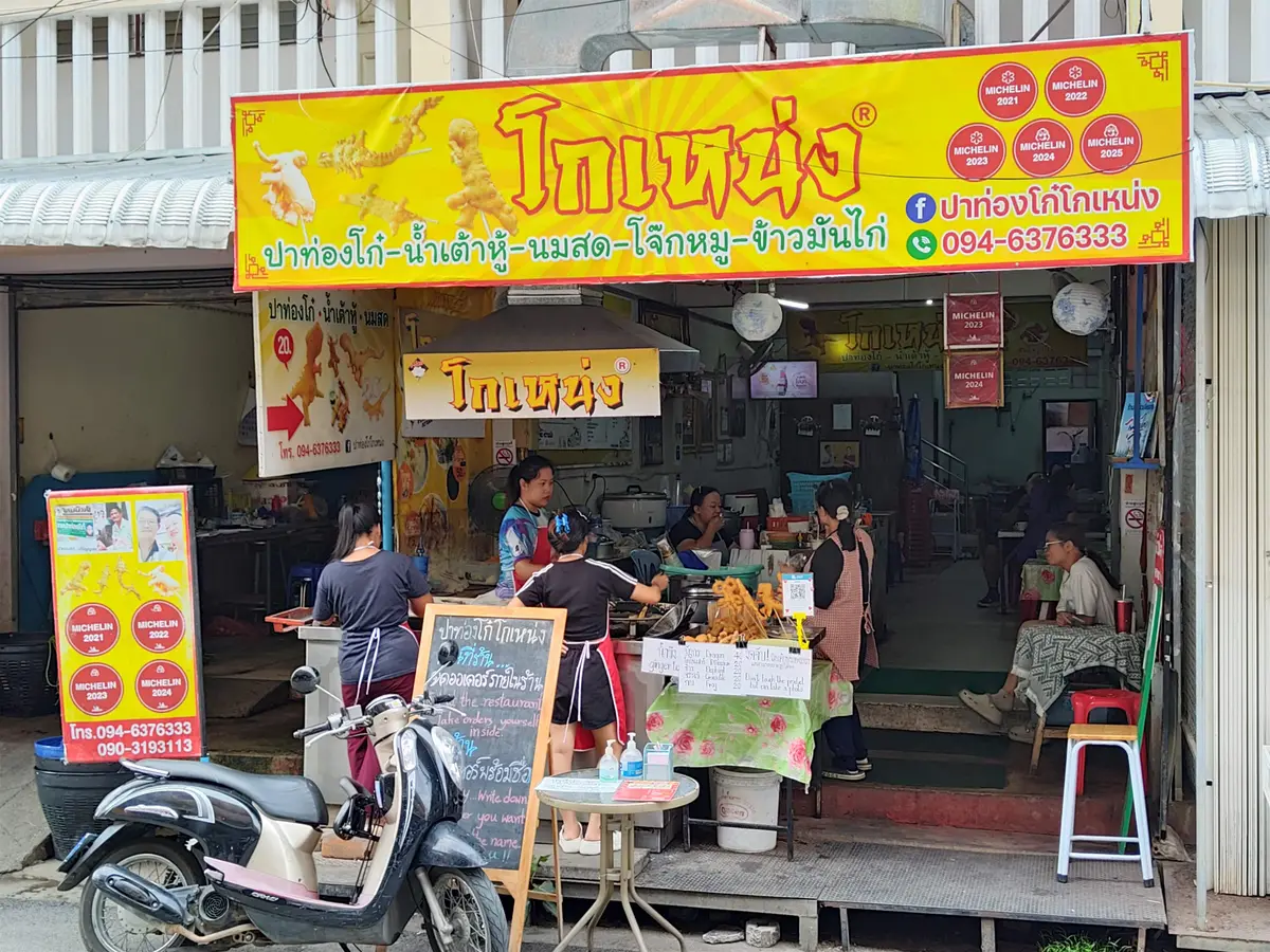 Street-side snack shop at the market with a large yellow sign in Thai and a motorbike parked in front.