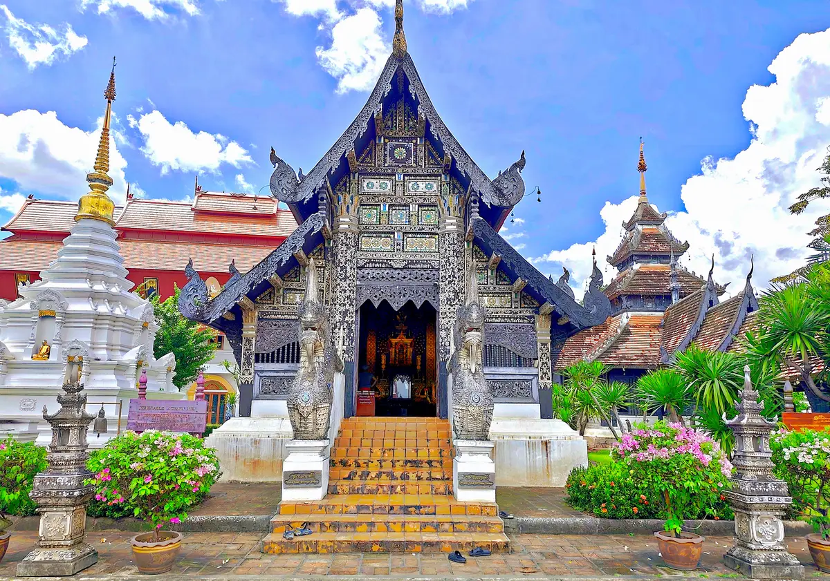 Ornate carved temple front with a steep roof and stone steps, with flowering plants in front