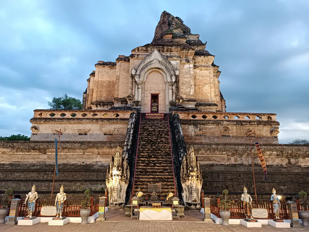 Brick chedi with a broken top rising above a stepped base and a central staircase flanked by statues