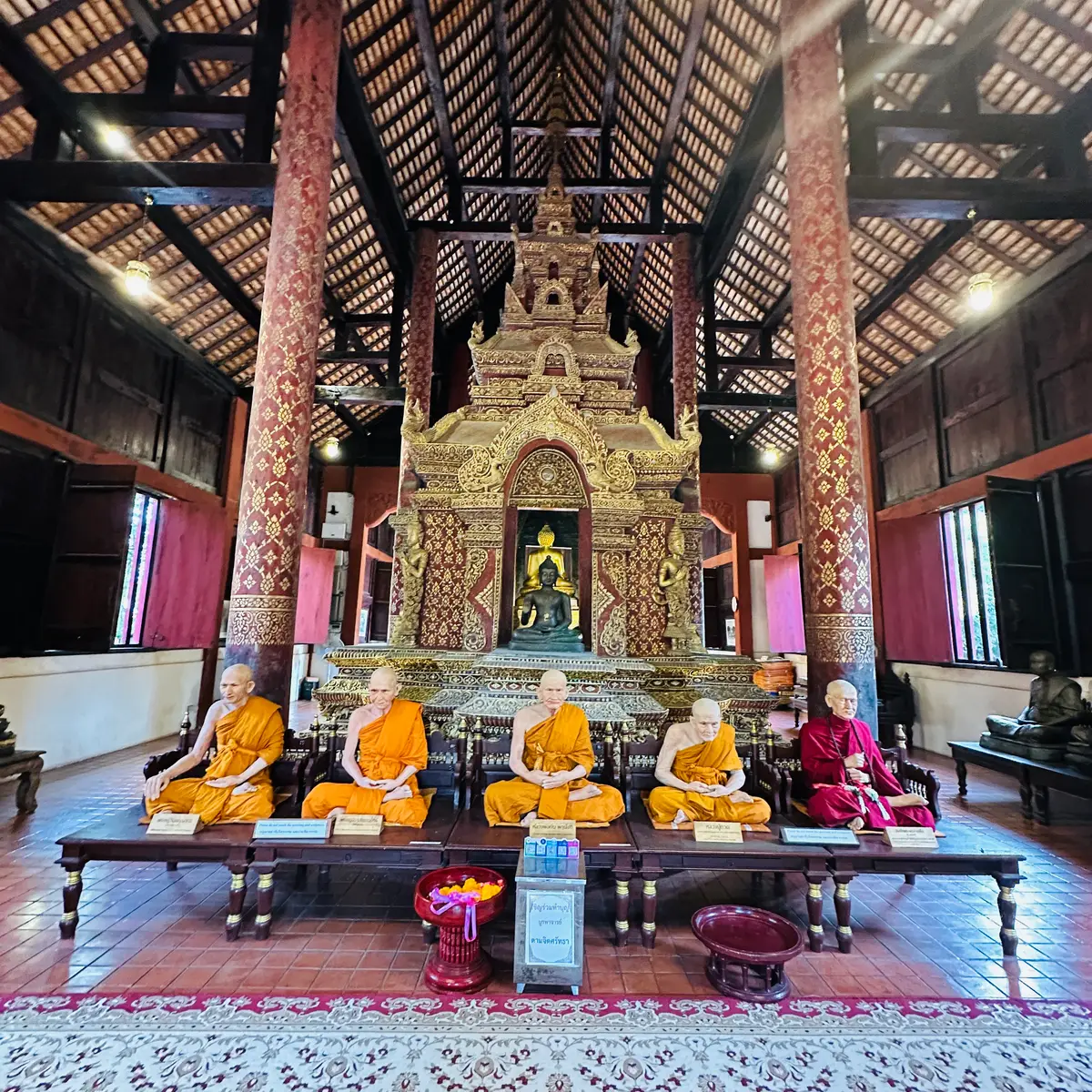 Life-size monk figures in orange robes seated at a table in front of a gilded shrine inside a hall