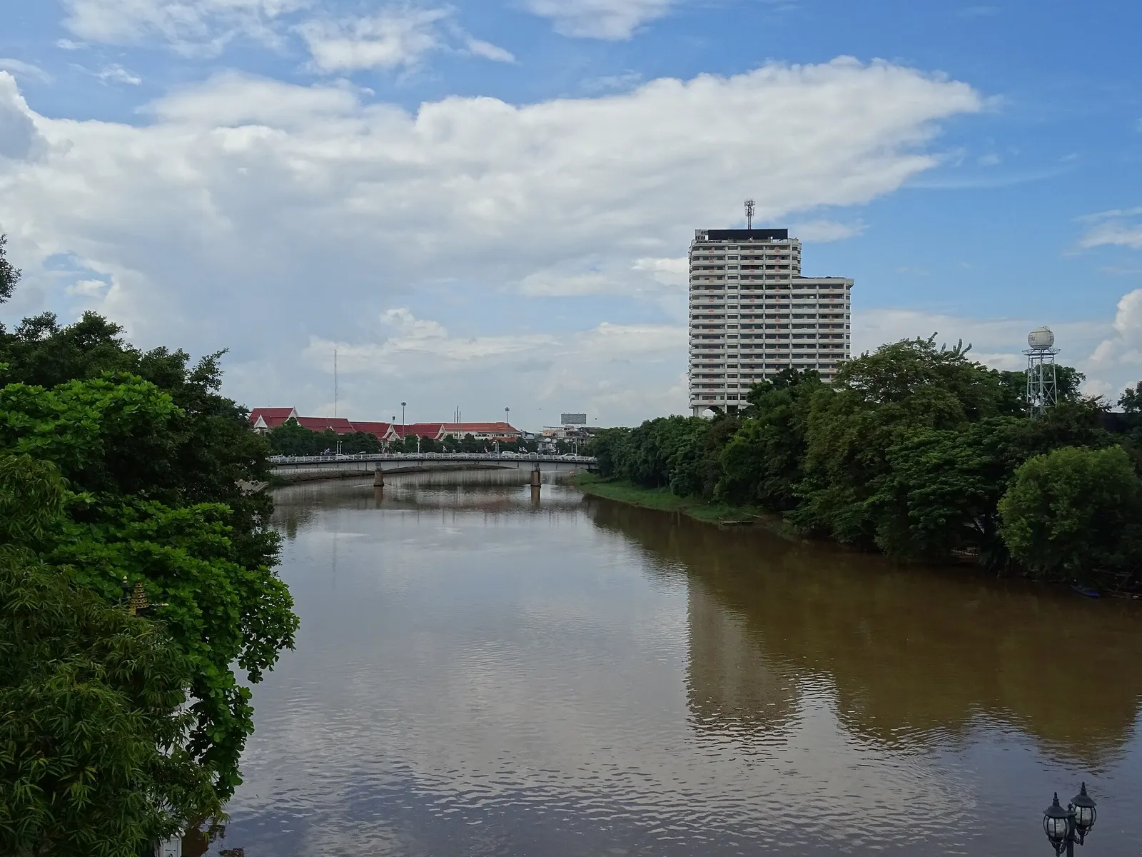 Brown river with a low bridge, a tall white building and green trees on the far bank